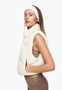 Pufferväst i off-white, ärmlös design med hög krage och vadderad mönstring. Bärs över en brun sportig topp, vilket visar en avslappnad passform.
