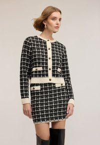 Černobílé šaty s kostkovaným vzorem pepito a k nim ladící krátký kardigan. Má knoflíky, kapsy a žebrované manžety. Elegantní textura látky.