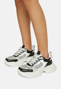 Sneaker chunky caratterizzata da un mix di materiali bianchi, neri e grigi, pannelli in rete, suole spesse e lacci prominenti. Design arrotondato e sovradimensionato.