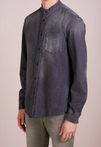 Camisa de mezclilla gris oscuro con cuello mao, mangas largas, cierre frontal de botones y un bolsillo en el pecho; acabado texturizado y ligeramente desgastado.