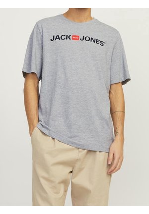 T-shirt gris à manches courtes avec logo noir "JACK & JONES" sur la poitrine. Le tissu semble doux. La main du modèle repose dans la poche d'un pantalon beige.