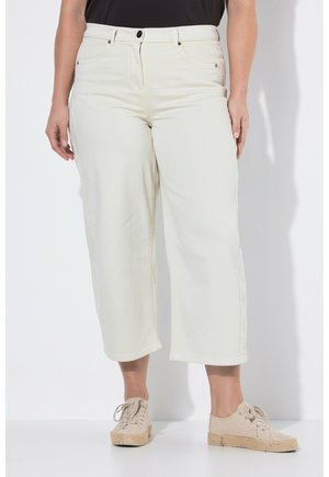 Donna che indossa pantaloni bianchi a vita alta, ampi e corti, abbinati a scarpe da ginnastica espadrillas beige e un top nero, in piedi davanti a uno sfondo bianco.