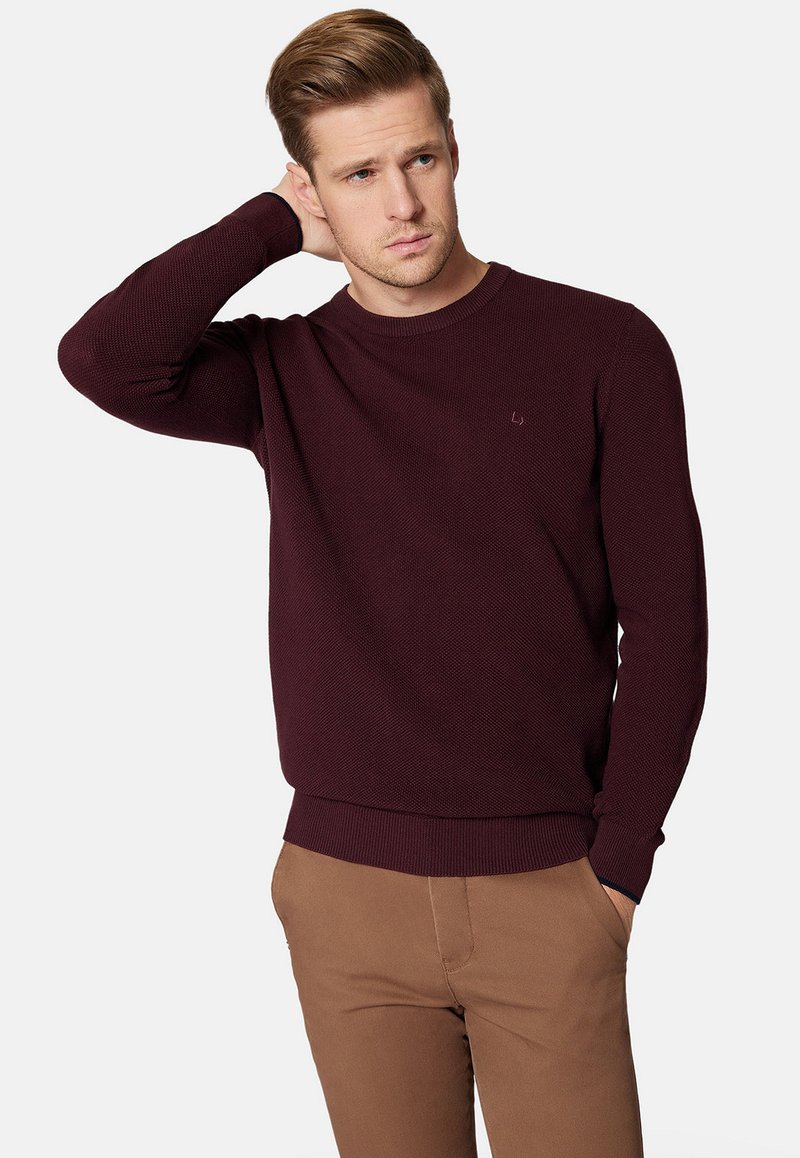 Bordowy sweter dzianinowy z okrągłym dekoltem i ściągaczami przy mankietach, w połączeniu z brązowymi spodniami. Posiada małe logo na klatce piersiowej.