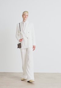 Blazer bianco doppiopetto con bottoni dorati, abbinato a pantaloni bianchi coordinati. Accessoriato con una piccola borsa scura. Texture liscia.