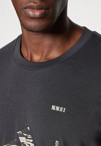 Camiseta de algodón en gris oscuro con cuello redondo, que presenta un texto gráfico blanco y un detalle de patrón texturizado en la esquina inferior.
