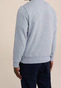 Man som bär en ljusgrå sweatshirt med långa ärmar och mörka byxor, sedd bakifrån mot en enfärgad beige bakgrund.