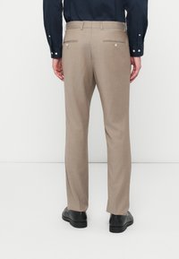 Ανοιχτό καφέ ραμμένα παντελόνια με λεία υφή, πτυχές και δύο πίσω τσέπες με φερμουάρ. Συνδυασμένα με μαύρα παπούτσια.