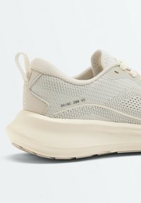 Vue de côté d'une basket beige tricotée avec une semelle épaisse crème et une languette au talon. Un petit texte "OY/AC 200 V2" est visible.