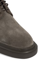 Chaussure en cuir suédé marron avec un bout rond et une épaisse semelle en caoutchouc noir sur fond blanc, montrant l'avant et une partie de la zone de laçage.