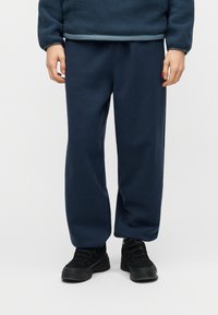 Άτομο που φοράει μπλε-σκούρο παντελόνι fleece, μαύρα παπούτσια με κορδόνια και μπλε-σκούρο φλις φούτερ, στέκεται μπροστά σε απλό φόντο.