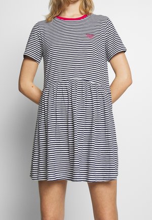 Kobieta ubrana w granatowo-białą sukienkę w paski z krótkim rękawem, marszczoną spódnicą i czerwonym wykończeniem przy dekolcie, stojąca z rękami za plecami.