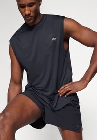 Black athletic tank top with sleeveless design and textured fabric, paired with black shorts featuring a side slit. Logo displayed on chest.