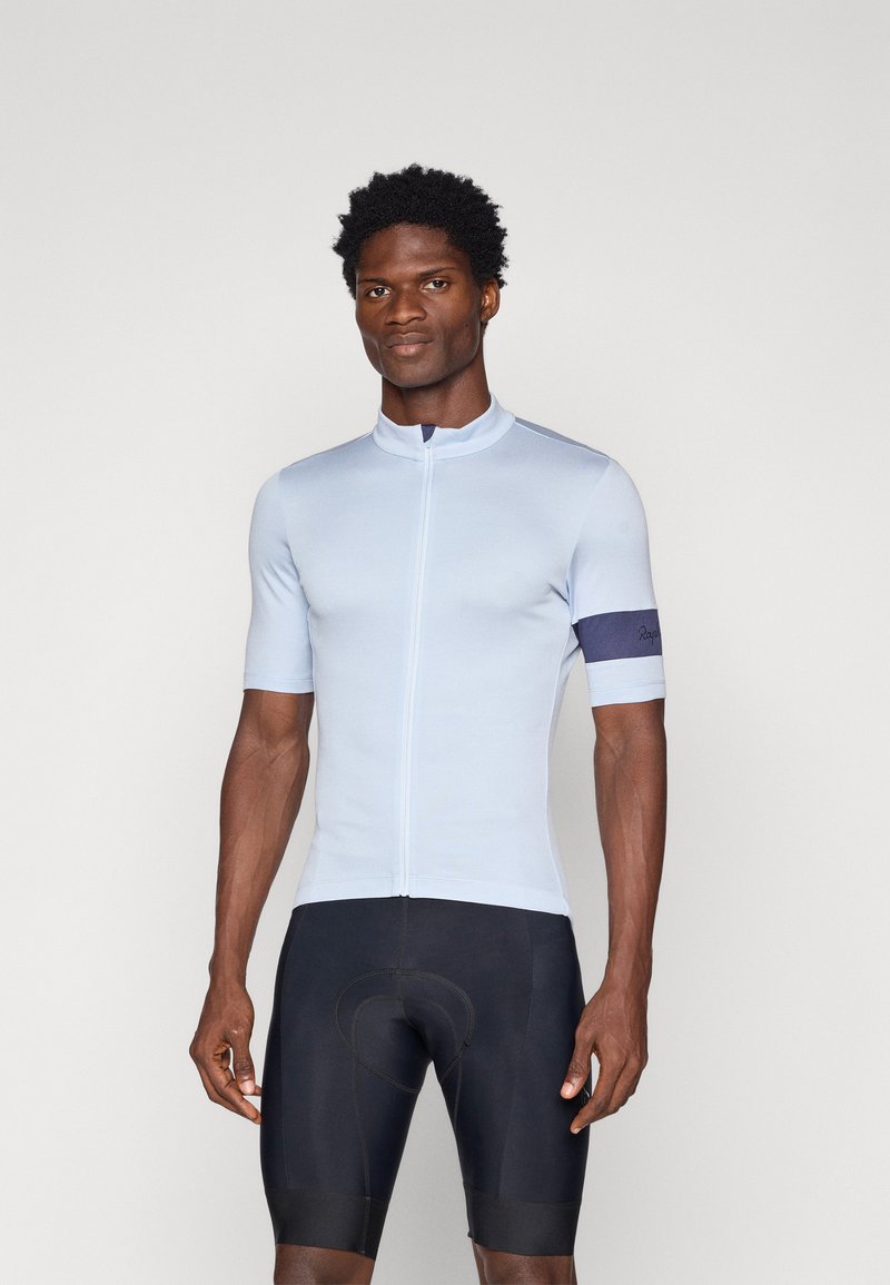 Tricou de ciclism bleu deschis, cu mâneci scurte, guler înalt, fermoar frontal și bandă de mână bleumarin închis; purtat cu pantaloni scurți negri de ciclism.