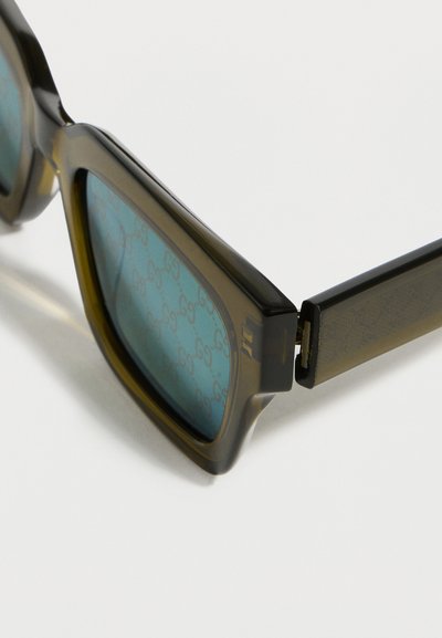 Gros plan sur des lunettes de soleil carrées teintées de vert avec une monture translucide vert olive foncé et un motif monogramme subtil sur les verres et les branches.