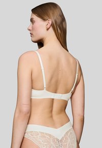 Krémová podprsenka s hladkým materiálem, nastavitelnými ramínky a zapínáním na háček a oko na zadní straně. Krajkový detail na odpovídajících kalhotkách.