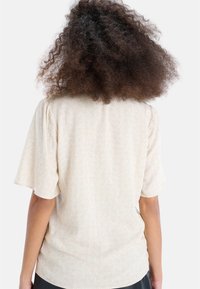 Světle béžová blůza s krátkými volánkovými rukávy a jemným vzorem listů. Měkká látka s hladkým povrchem, volný střih.