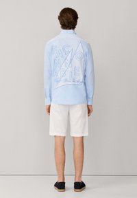 Man draagt lichtblauw shirt met lange mouwen met grote omranderde tekst en abstracte vorm op de achterkant, witte korte broek en zwarte instapschoenen, staand.