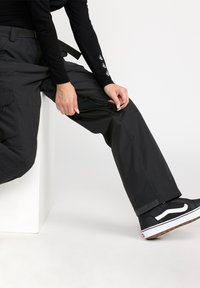 Sorte vandtætte bukser med en lynlåslomme på låret, afslappet pasform og et bælte. Båret med sorte og hvide sneakers.