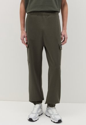 Pantalones cargo color verde oliva con cintura elástica, bolsillos laterales y puños elásticos, combinados con zapatillas blancas. Textura de tela suave.