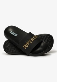 Black slide sandals with a wide strap featuring the word "SUPERDRY" in gold lettering. Textured black rubber sole and smooth upper material.