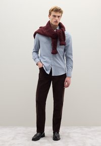 Camicia azzurro chiaro con bottoni, pantaloni di velluto a coste marrone scuro, sciarpa lavorata a maglia color bordeaux elegantemente drappeggiata intorno al collo, scarpe nere, sfondo semplice.