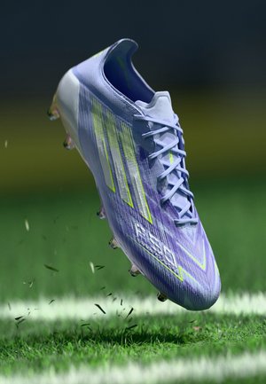 Light purple soccer cleat with neon yellow stripes and studs, lifting from green grass on a marked field.