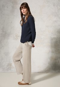 Marineblauwe gebreide trui over een witte blouse met kraag, gecombineerd met lichtbeige wijde broek en beige sneakers met bruine accenten. Glad textuur.