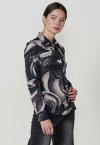 Blusa a maniche lunghe con bottoni, di colore nero, decorata con motivi astratti grigi e bianchi a vortice. Realizzata in tessuto morbido con colletto classico.