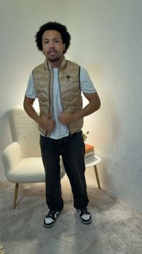 Άνδρας με μπεζ επενδεδυμένο γιλέκο πάνω από λευκό πουκάμισο, διορθώνει το γιλέκο του, στέκεται σε χαλί κοντά σε λευκή πολυθρόνα και μικρό τραπέζι με βιβλία και φυτό.