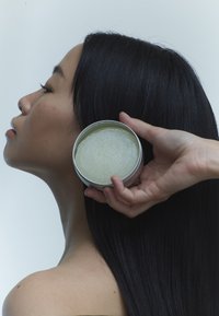 Boîte ronde en étain argenté contenant une substance texturée de couleur vert clair à côté du profil d'une personne aux longs cheveux noirs, sur un fond pâle.