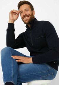 Marineblaues Zip-Hoodie mit hohem Kragen und Brusttasche, kombiniert mit hellblauen Jeans. Glatter Stoff mit gerippten Bündchen.