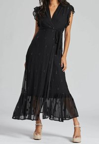 Schwarzes Wickelkleid mit Rüschensleeves, das einen transparenten Rock, strukturierten Stoff und dekorative goldene Akzente aufweist. Langer, fließender Schnitt.