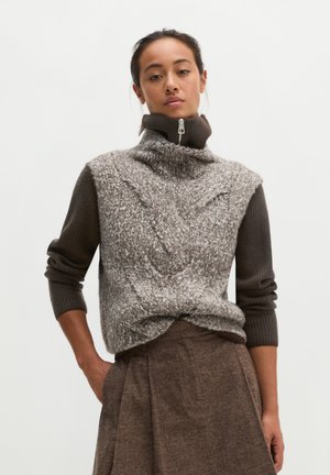 Vrouw draagt een grijs gestructureerd gebreid vest over een bruine geribde coltrui en bruine hoge taille broek met zakken.
