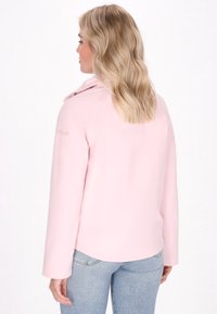 Ženska z valovitimi blond lasmi nosi roza jakno in modre kavbojke, vidna od zadaj, kar prikazuje ovratnik in stil jakne.