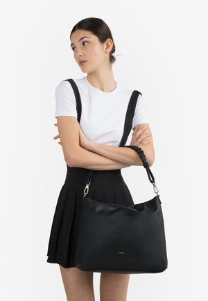 Donna con t-shirt bianca e gonna nera che tiene una grande borsa nera con manico intrecciato, guardando di lato.