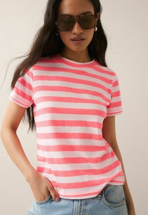 Woman wearing tortoiseshell sunglasses, gold hoop earrings, pink and white striped T-shirt, and light blue jeans with one hand in pocket.