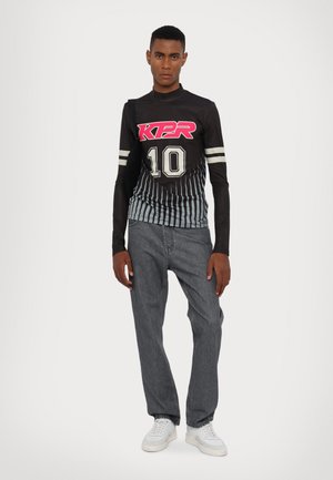Sort langærmet top med pink "KPR" logo og hvide striber, med lodret ribbet tekstur, parret med grå lige ben jeans og hvide sneakers.