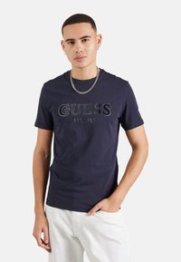 Camiseta de algodón azul marino con el logo embossado "GUESS EST. 1981". Ajuste clásico, mangas cortas y diseño de cuello redondo. Llevada con pantalones blancos.