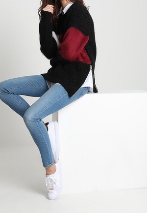 Donna seduta su un blocco bianco che indossa un maglione nero e rosso, jeans attillati azzurri e scarpe da ginnastica bianche.