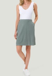 Femme portant un top sans manches blanc et une jupe à motifs de longueur genou avec des poches, se tenant avec une main dans la poche et un pied croisé.