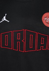 Czarny trykot z wyraźnym czerwonym logo "JORDAN" grubą czcionką, białym haftowanym logo Jumpman oraz okrągłą metką z informacjami o marce.