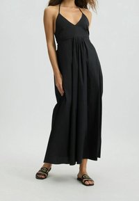 Musta maxi-mekko, jossa on V-pääntie, nyörilliset olkahihnat ja kerrottu vyötärö. Vaatteen kangas on sileä ja laskeutuva. Yhdistetty mustiin liukuviin sandaaleihin, joissa on kultaiset yksityiskohdat.