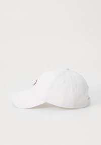 Biela čiapka vyrobená z bavlny, s zakriveným šiltom, nastaviteľným popruhom a malým viacfarebným logom na prednej strane.