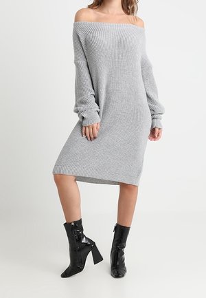 Femme portant une robe grise en maille à épaules dénudées et des bottes noires en cuir verni à talons mi-mollet, debout devant un fond blanc.