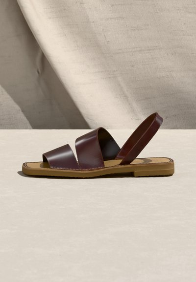 Καφέ δερμάτινο σανδάλι με χαμηλό τακούνι, δύο φαρδιές φάσες πάνω από το πόδι και λουράκι στην πλάτη, τοποθετημένο πάνω σε ουδέτερο υφασμάτινο φόντο.