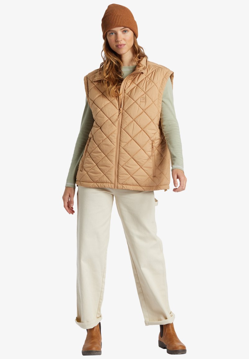 Gilet trapuntato beige con colletto alto, zip frontale e due tasche laterali, abbinato a una camicia a maniche lunghe verde chiaro e pantaloni crema.