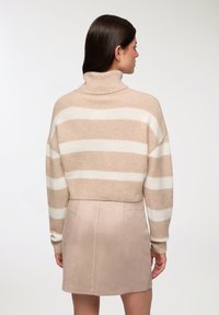 Kobieta zwrócona plecami w beżowo-białym paskowanym swetrze z golfem i beżowej spódnicy na tle jednolitej białej ściany.