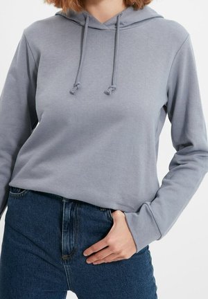Felpa grigia in morbido tessuto, con tasca frontale, cappuccio regolabile e maniche lunghe. Abbinata a jeans a vita alta.