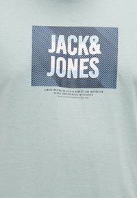 Ανοιχτό μπλε μπλουζάκι με ορθογώνιο ναυτικό γραφικό και λευκό κείμενο "JACK&JONES" και επιπλέον μικρή εκτύπωση παρακάτω σε μαύρο χρώμα.
