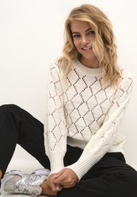 Weißer gestrickter Pullover mit Rautenmuster, Lückenmuster und gerippten Bündchen, kombiniert mit schwarzen Hosen und grauen Sneakers. Model sitzt und lächelt.